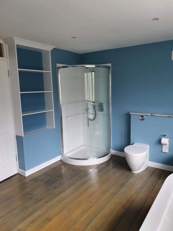 Picture of a corner shower unit besides a toilet in a bathroom that has been painted with a dull blue emulsion. The floorboards have been sanded and varnished to a classic finish.