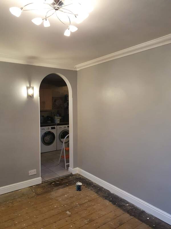 Picture a dining room leading into a utility room. The walls of the dining room are painted in light grey and the skirting boards have been painted in white gloss. There is white coving around the ceiling to wall junction and the original floorboards are exposed. 