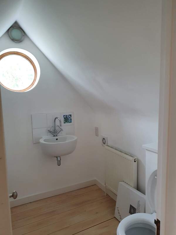 Picture an attic room en-suite. There is a circular window on the wall opposite the door into the room letting natural daylight in. The walls to the room has been finished in a white matt emulsion. The floor is a timber laminate finish