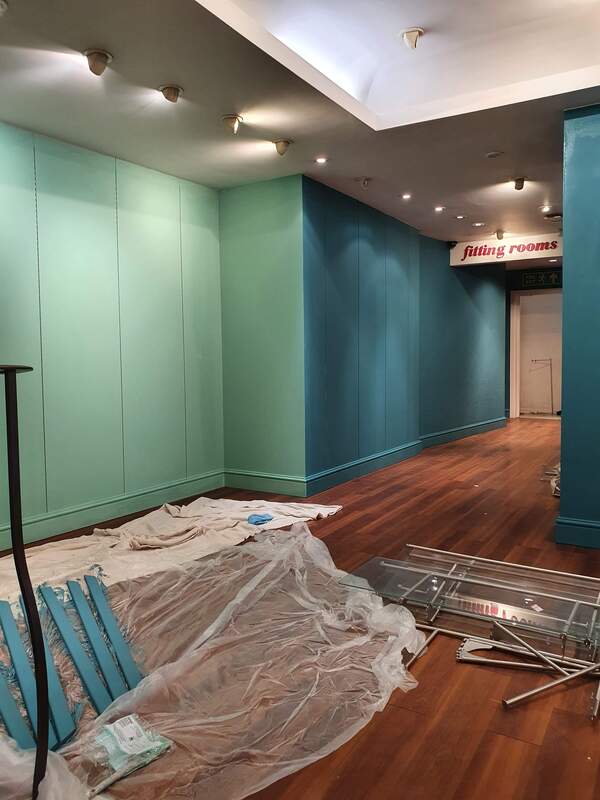 Picture of a retail shop under going redecoration. The floor has a dark oak laminate, which is contrasted by a teal and light blue emulsion on the walls. The fitting rooms are visible to the rear of the shop. 