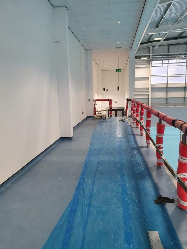 Picture of an internal mezzanine walkway situated within a school sports hall. The sports hall is under going refurbishment and the walls are painted in white emulsion. The blue hardwearing flooring is covered with a dust sheet to protect from damage. 