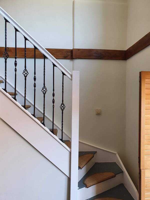 Picture of the bottom of a domestic staircase. The metal railings on the stairs are painted in black gloss and the handrail is a contrasting white gloss. The wall behind is painted in white emulsion with a dark oak beam running horizontally around the centre of the wall. 